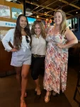 Tres mujeres posan juntas en un restaurante informal. La de la izquierda lleva una blusa blanca y pantalones cortos azules, la del centro una camisa a rayas y pantalones negros, y la de la derecha un vestido floral.