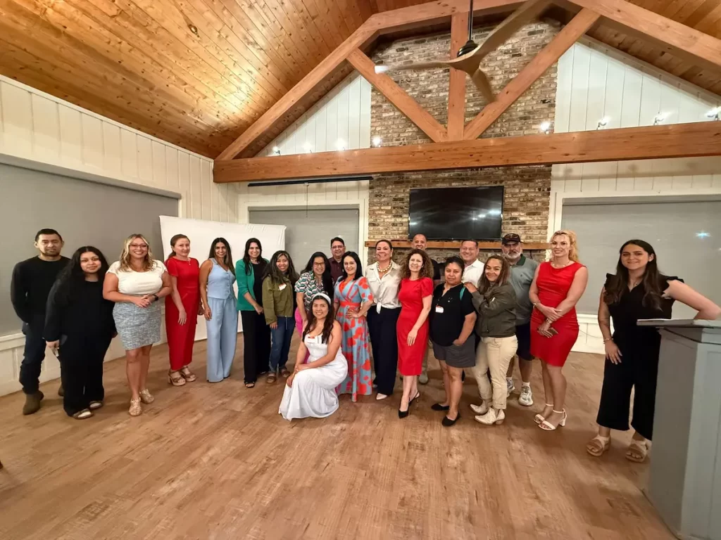 A group of 20 people poses and smiles together in a spacious room with wooden floors, a tall ceiling with wooden beams, and a stone wall with a TV. Some are dressed formally in dresses and suits.