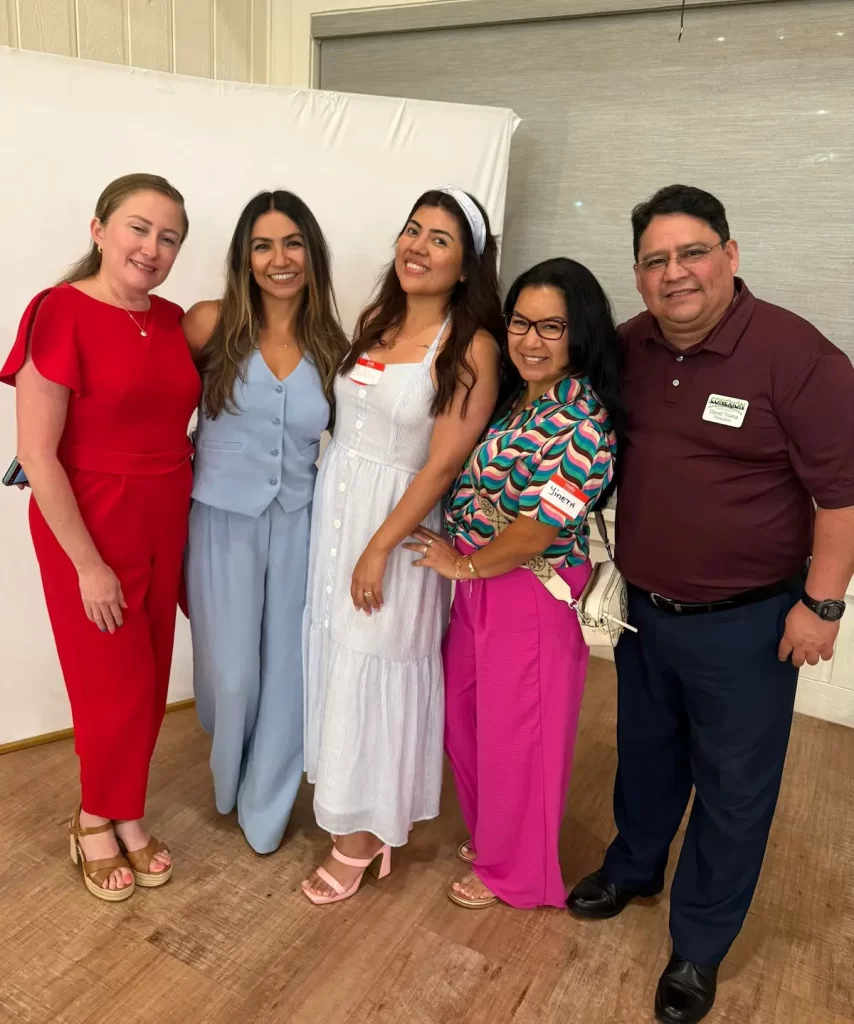 Five people stand together smiling for a group photo indoors, each wearing name tags. They are dressed in colorful, semi-formal outfits and pose closely, with a neutral background behind them.
