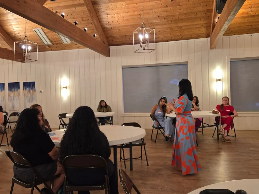 A woman in a colorful dress stands speaking to a group of people seated at round tables in a wooden-ceilinged room with white paneled walls and hanging lights.