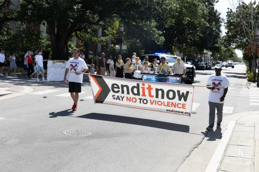 Dos personas sostienen una pancarta en la que se lee "enditnow SAY NO TO VIOLENCE" mientras encabezan una marcha contra la violencia y la trata de seres humanos; una banda de música y un coche de policía les siguen, mientras los espectadores permanecen de pie en la soleada acera.