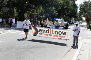 Dos personas sostienen una pancarta en la que se lee "enditnow SAY NO TO VIOLENCE", encabezando un grupo que marcha por una calle de la ciudad para concienciar sobre la violencia doméstica; al fondo se ve un coche de policía, una banda y transeúntes.