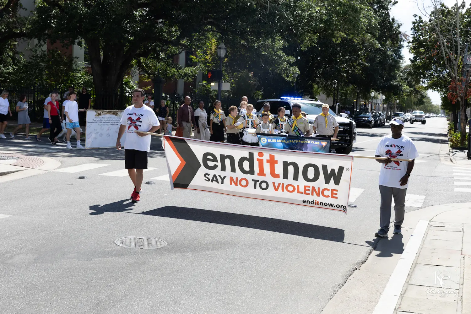 Dos personas sostienen una pancarta en la que se lee "enditnow SAY NO TO VIOLENCE", encabezando un grupo que marcha por una calle de la ciudad para concienciar sobre la violencia doméstica; al fondo se ve un coche de policía, una banda y transeúntes.