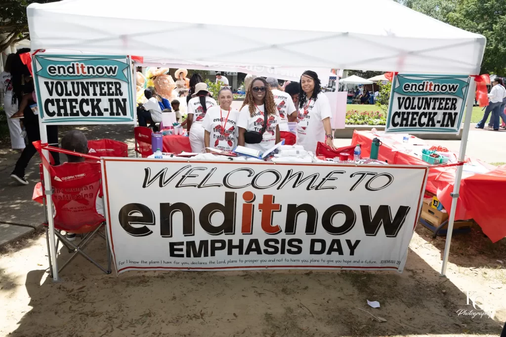 Tres personas de pie detrás de una mesa en un puesto de un evento al aire libre con una gran pancarta "Welcome to enditnow Emphasis Day" (Bienvenido al Día de Énfasis en los Derechos Humanos), donde hay carteles que abordan temas como el acoso escolar y la violencia. El stand está rodeado de material del evento y un cartel de "Registro de voluntarios".