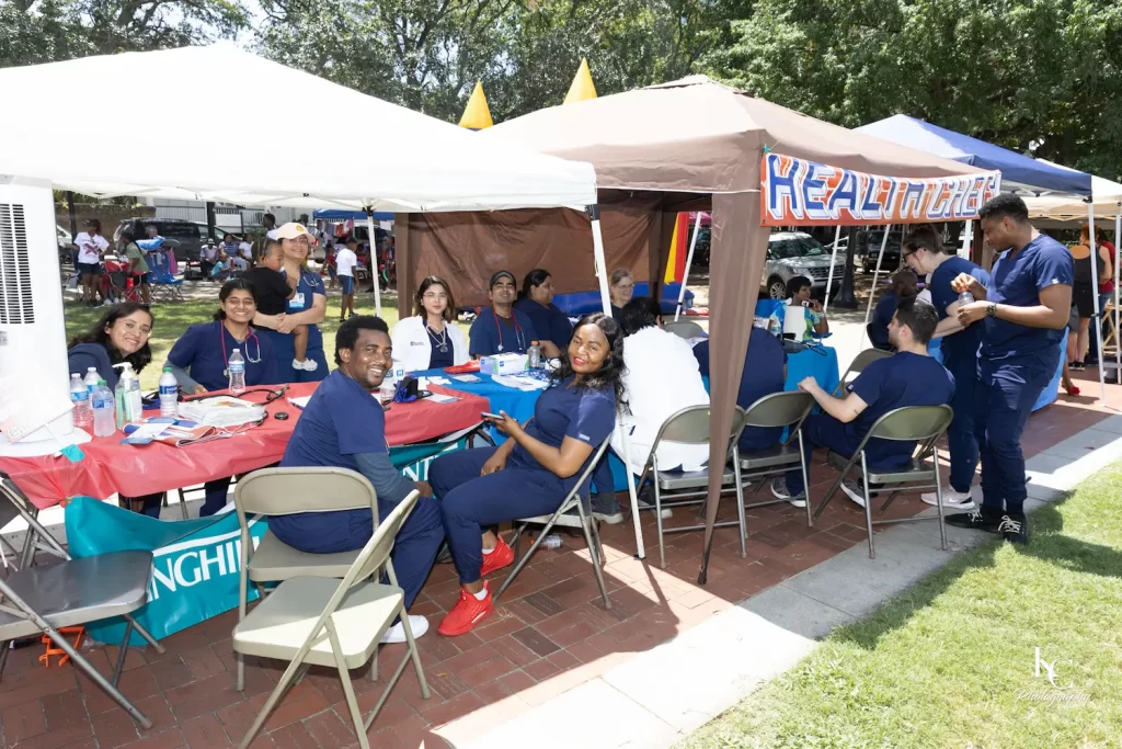 Un grupo de personas vestidas con batas azules se sientan y permanecen de pie bajo carpas al aire libre con la etiqueta "SALUD", sonriendo y hablando en las mesas durante un soleado acto comunitario en un parque, compartiendo recursos sobre la prevención del acoso escolar.