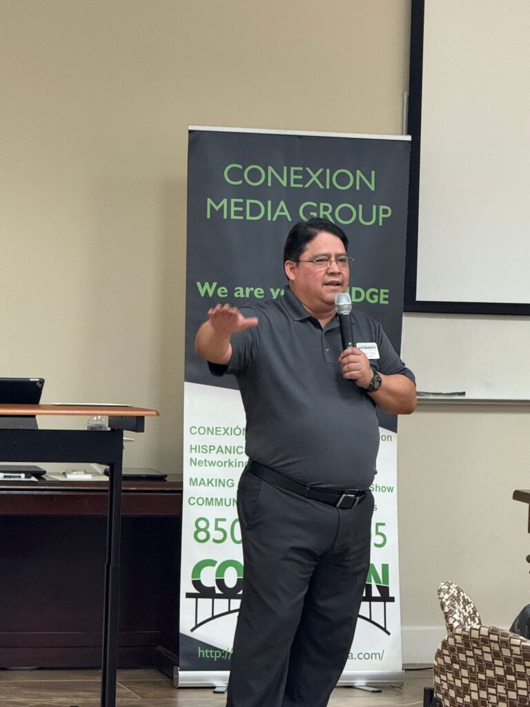 A man in a gray shirt speaks into a microphone while gesturing with his hand. He stands in front of a "Conexion Media Group" banner during an indoor event or presentation.