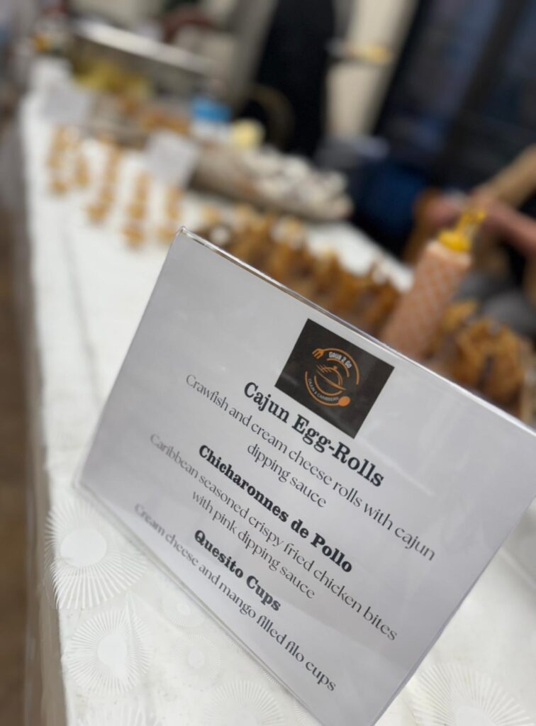 A close-up of a menu card on a table displaying food options including Cajun Egg-Rolls, Chicharrones de Pollo, and Quesito Cups. Food platters are visible in the blurred background.