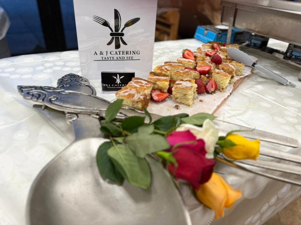 A catering table displays sliced pastries with strawberries, a serving spoon and fork, three roses (red, yellow, and pink), and an A & J Catering sign on a white tablecloth.