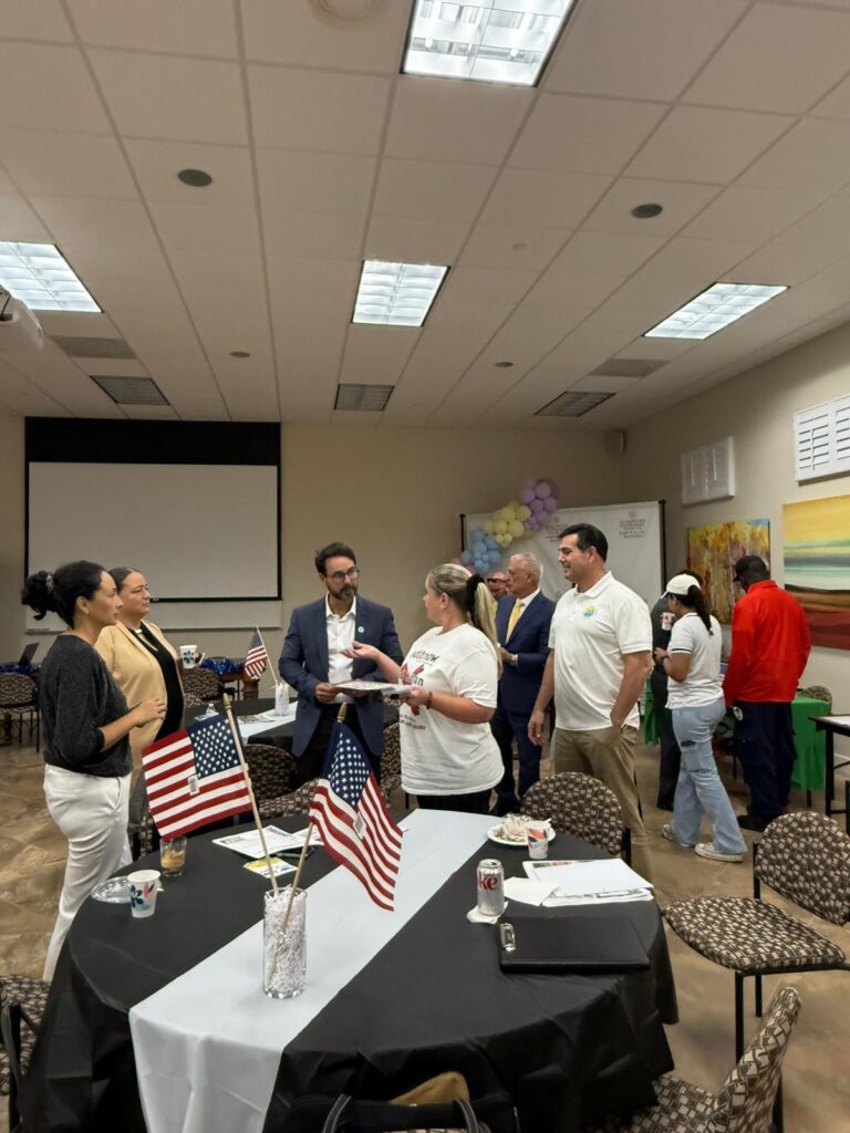 A group of people gather and converse in a decorated room with American flags on a table. Some stand in small groups, while others walk around. The atmosphere appears friendly and casual.