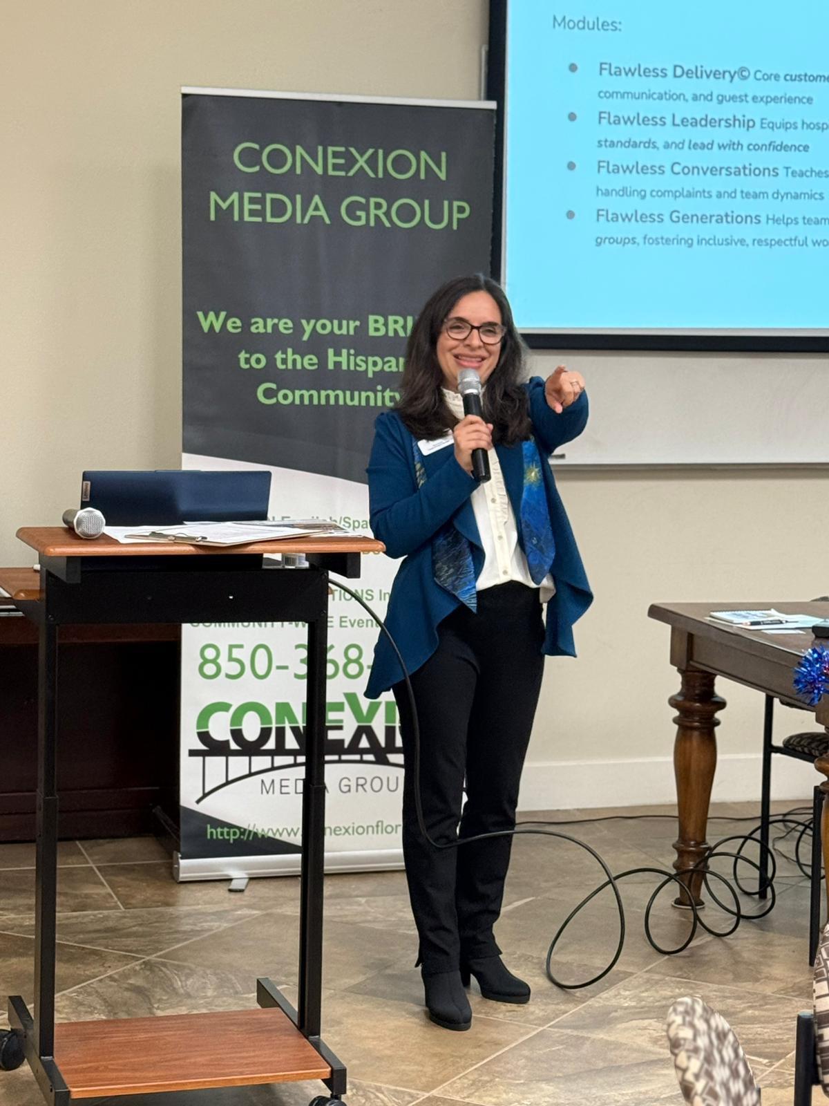 A woman in business attire smiles and points toward the camera while speaking at a podium. Behind her are a screen displaying a presentation and a sign reading "CONEXION MEDIA GROUP.