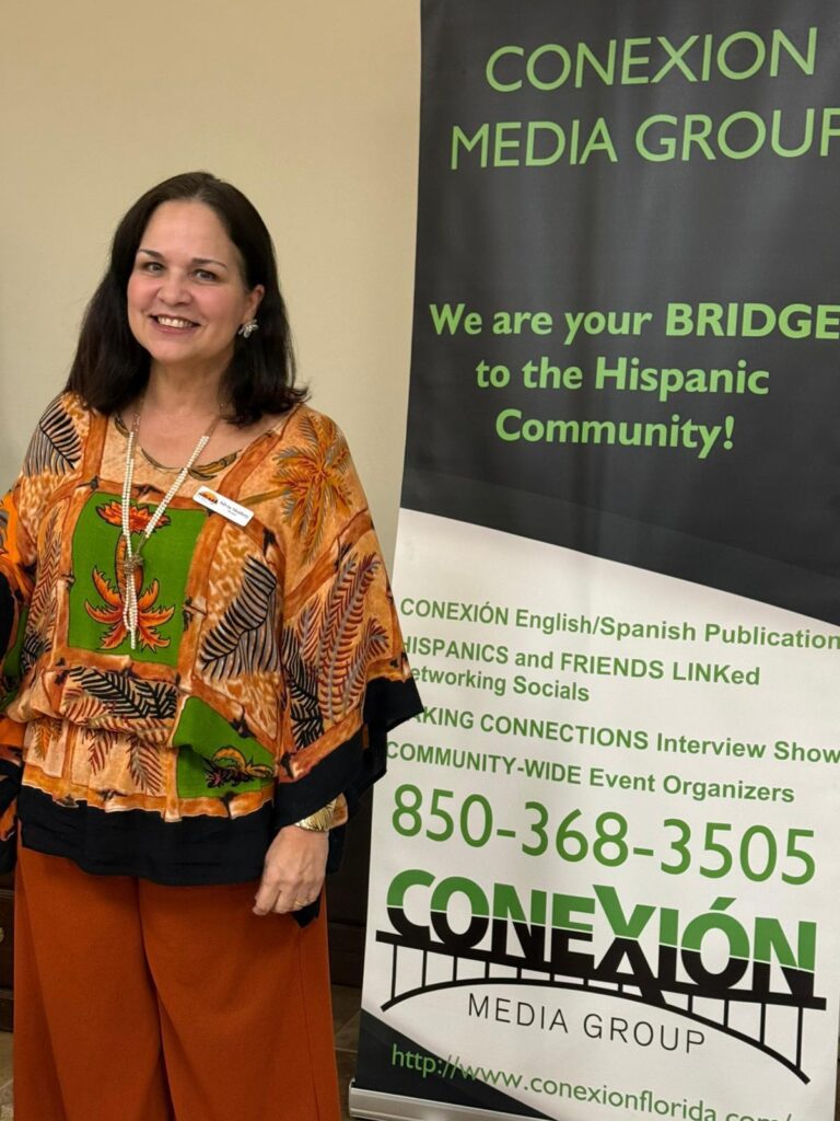 A smiling woman in an orange and patterned blouse stands next to a banner for CONEXIÓN Media Group, which offers services to the Hispanic community, including publications and networking events.
