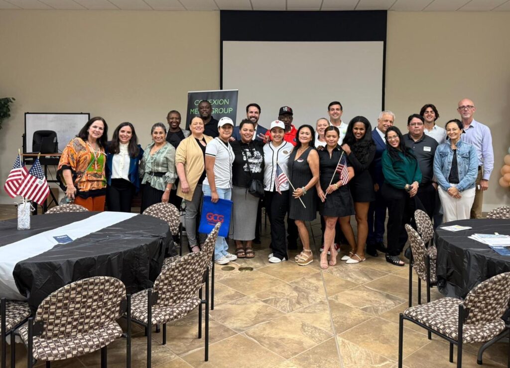 A diverse group of people pose together indoors at an event, smiling in front of a projection screen and a sign. Some hold small American flags. Tables with black tablecloths and chairs are arranged in the foreground.