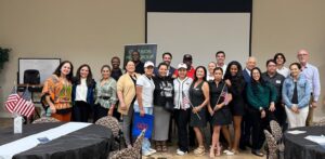 A diverse group of about 25 people poses together indoors, smiling at the camera. Some hold small American flags. They stand in front of a table and a white projection screen in a well-lit room, suggesting a community or networking event.