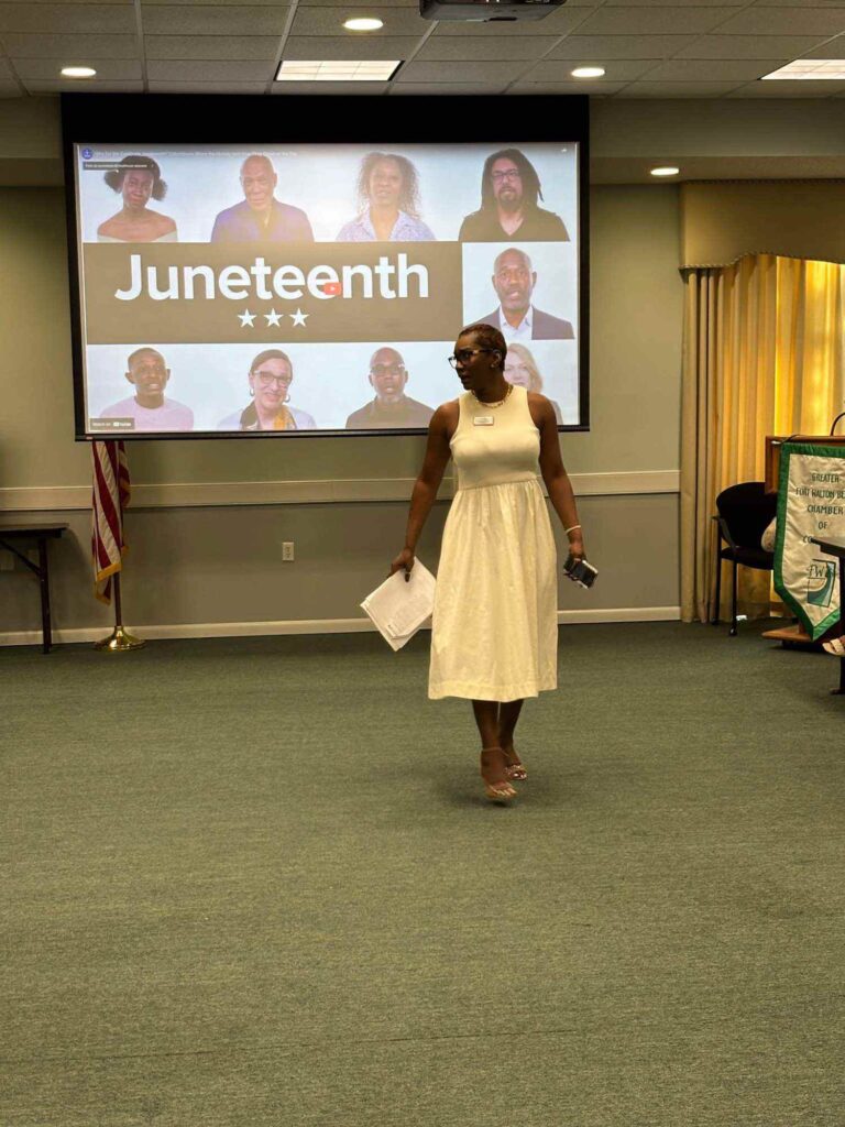 A woman in a white dress stands in a room holding papers, with a Juneteenth presentation displayed on a screen behind her showing photos of diverse people. An American flag and yellow curtains are in the background.
