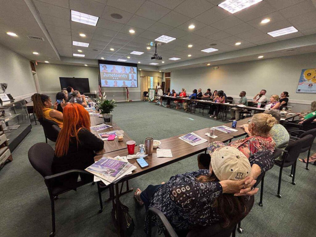 A group of people sit around a large conference table watching a presentation about Juneteenth displayed on a screen at the front of the room.