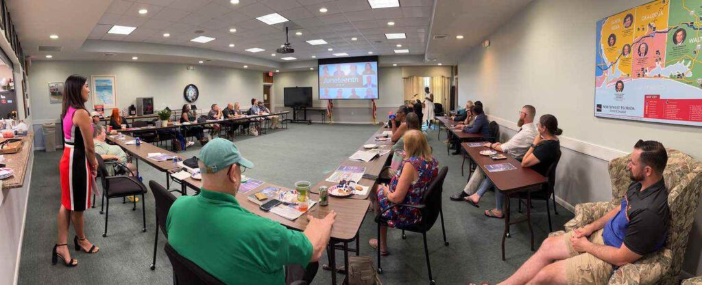 A group of people sits around U-shaped tables in a conference room, watching a presentation on a large screen. A woman stands at the front speaking. A map is displayed on the wall to the right.