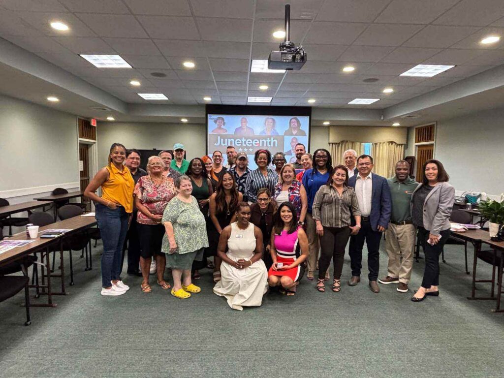 A diverse group of people poses and smiles for a photo in a conference room, with a screen in the background displaying "Juneteenth." Tables and chairs are visible along the sides of the room.