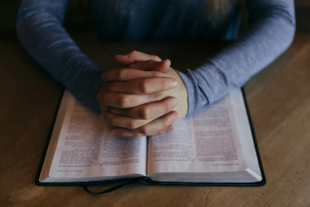 Una persona con camisa azul de manga larga se sienta ante una mesa de madera con las manos juntas en señal de oración sobre un libro abierto.
