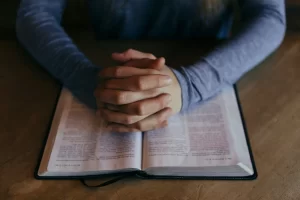 A person in a long-sleeve blue shirt sits at a table with hands clasped in prayer over an open Bible.