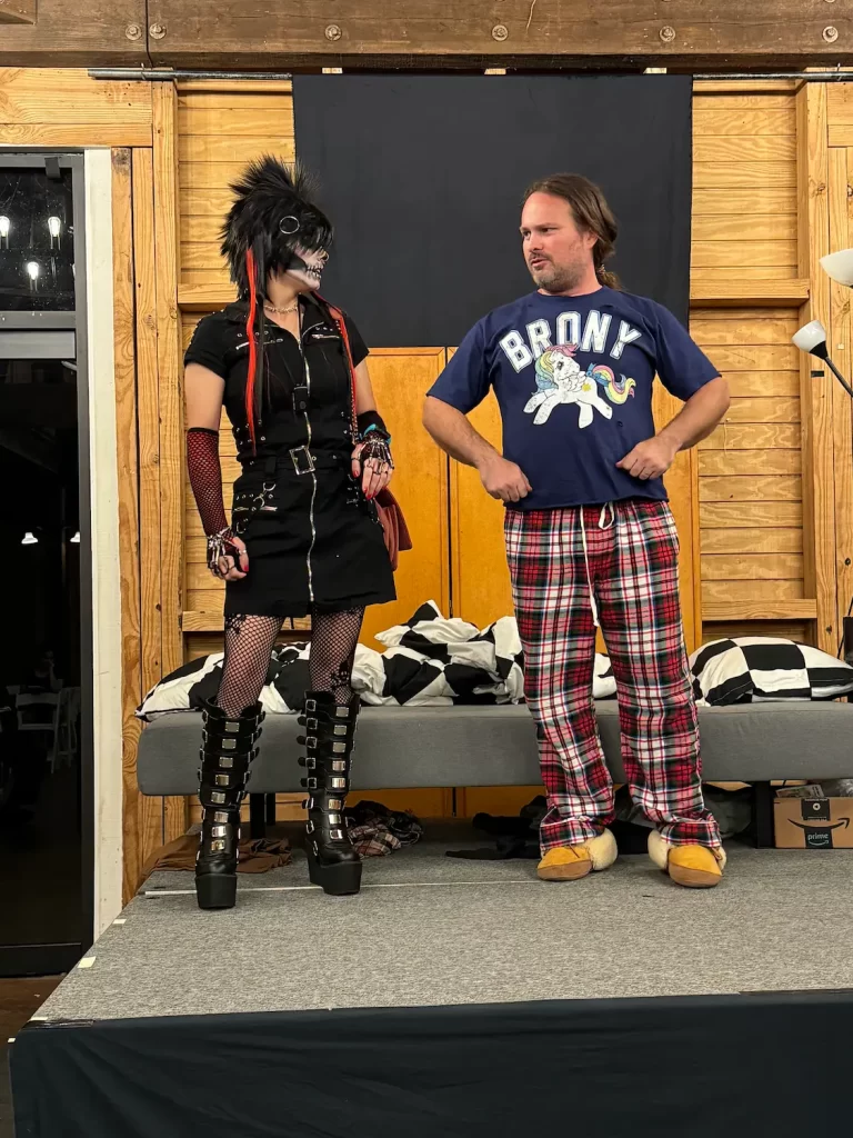 Two people stand on stage at the Eighth Annual Micro Theater Festival. The left wears gothic attire with platform boots, fishnets, gloves, and a spiked mask. The right rocks a "BRONY" T-shirt, plaid pajama pants, and animal paw slippers.