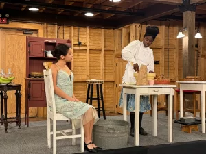Two women in period costumes perform on stage at the Eighth Annual Micro Theater Festival. One sits in a light blue dress, watching as the other, in a white blouse and patterned skirt, prepares something with kitchen props in a rustic wooden setting.