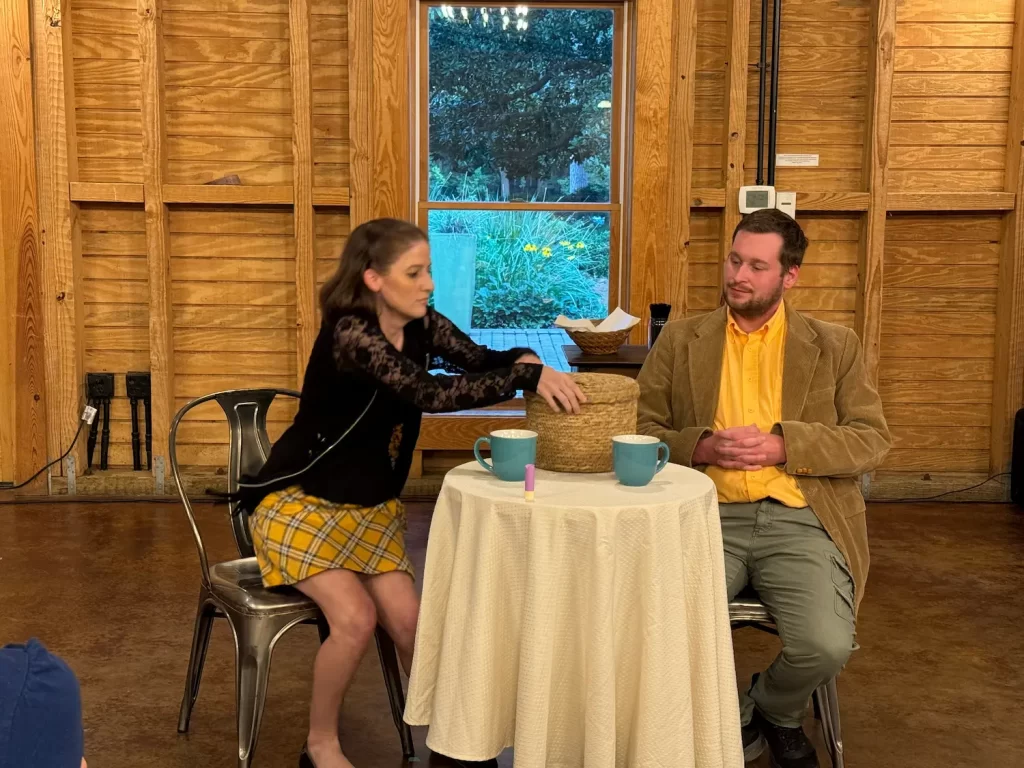 At the Eighth Annual Micro Theater Festival, a woman and man sit at a small round table with a white tablecloth, two mugs, and a basket. The woman reaches for the basket as greenery peeks through the wooden room’s window.