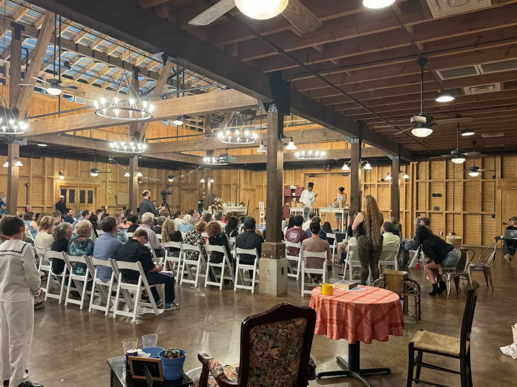 A large group of people sit on white chairs inside a rustic wooden hall, watching an auctioneer and assistants conducting an auction on a raised platform under bright chandeliers during the Eighth Annual Micro Theater Festival.
