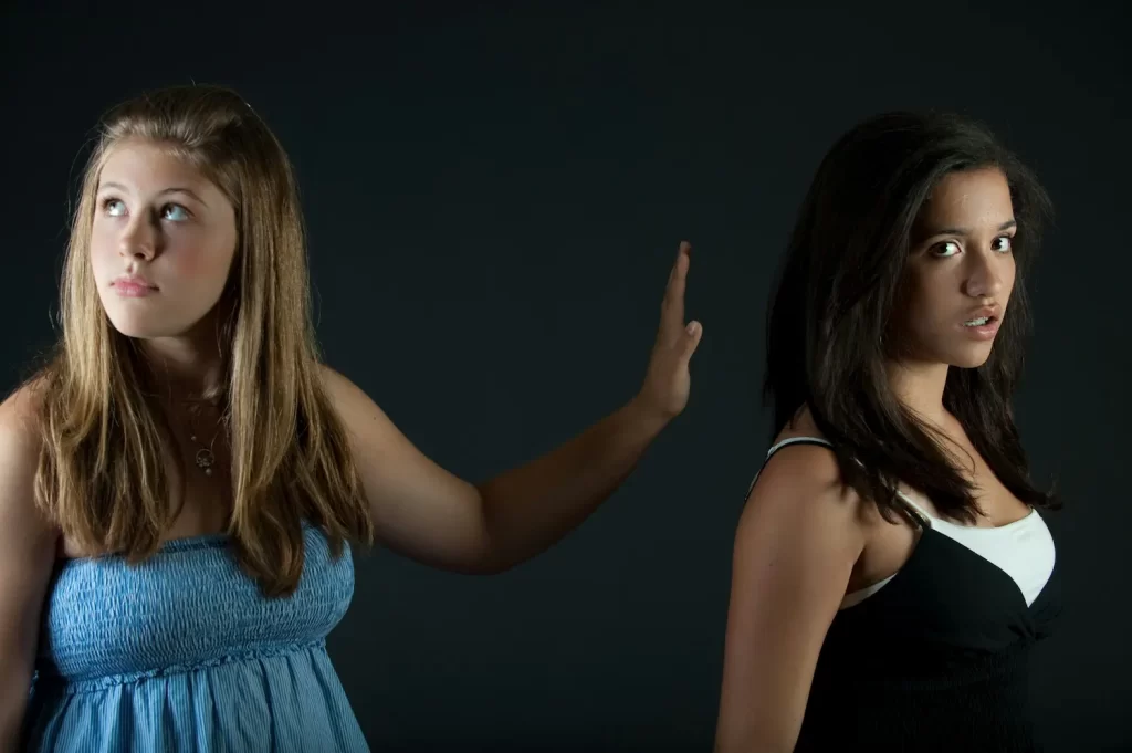 Two teenage girls stand against a dark background. One girl faces forward with her hand raised as if to stop the other, highlighting a moment of conflict; the other looks away, her expression distant—capturing the challenge of handling conflict.