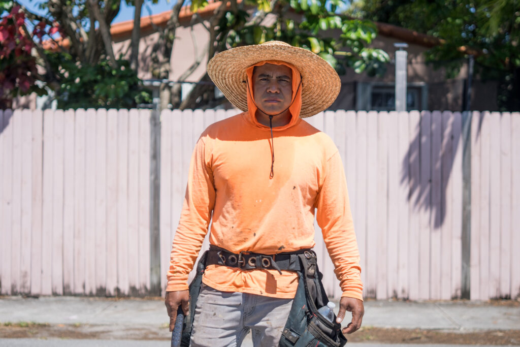 Una persona vestida con una camisa naranja de manga larga, un sombrero de paja de ala ancha y un cinturón de herramientas se encuentra en el exterior frente a una valla de madera de color rosa claro en un día soleado.