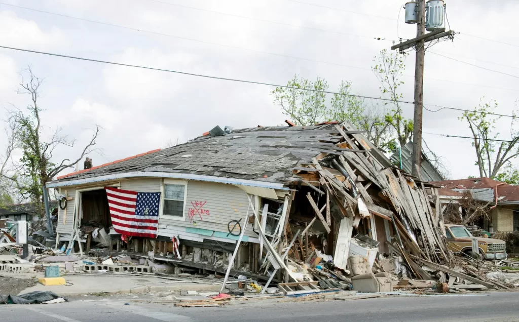 A severely damaged house with a collapsed roof and debris scattered around, likely impacted by hurricanes. An American flag hangs on the front, with power lines and a utility pole nearby. The surrounding trees show damage, highlighting disaster inequality.