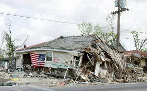 A severely damaged house with a collapsed roof and debris scattered around, likely impacted by hurricanes. An American flag hangs on the front, with power lines and a utility pole nearby. The surrounding trees show damage, highlighting disaster inequality.