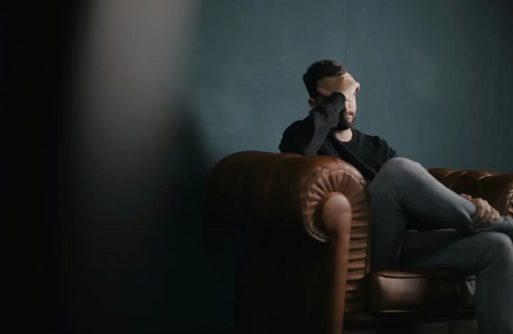 A man sits alone on a brown leather sofa with his legs crossed, resting his head on one hand, looking stressed or deep in thought—perhaps reflecting on mental health—against a dark green wall.