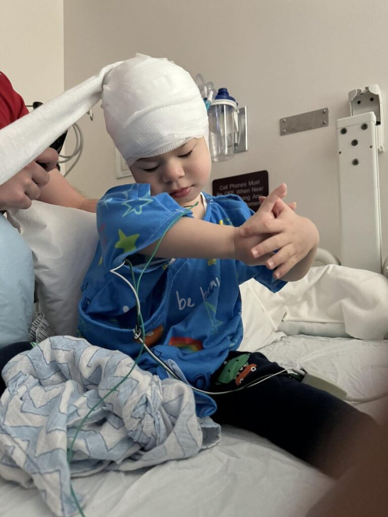 A young child with disabilities and a bandaged head sits on a hospital bed, wearing a blue hospital gown with colorful designs. Medical wires are attached to their body, while an adult assists from the side.
