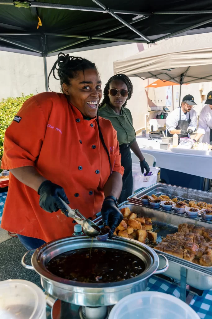 A smiling chef in an orange coat serves food at an outdoor market stall, using tongs to dish from a large pot. Trays of food are on the table, with another person and more vendors in the background.