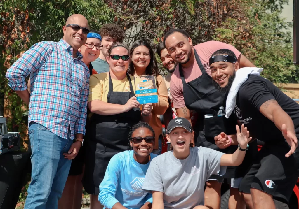 A group of smiling people, some wearing aprons, gather outdoors for a photo. Two people kneel in front, and one person holds a box of graham crackers. Trees and sunlight are visible in the background.