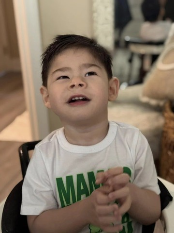 Un niño de pelo corto y oscuro que lleva una camisa blanca con letras verdes está sentado en el interior de una casa, mirando hacia arriba y sonriendo con las manos juntas. La escena, con una iluminación suave y un espejo de fondo, destaca la alegría y la inclusión de los niños con discapacidad.