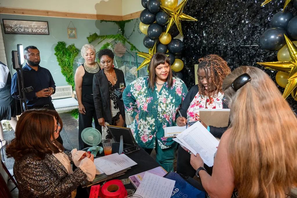 A group of people stand in line at a decorated check-in table with black and gold balloons and star decorations. Two women review paperwork while others wait and observe in a festive indoor setting.