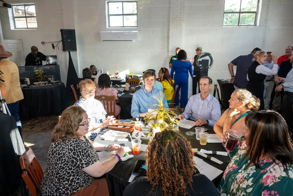 A group of people sit and stand around tables in a bright, spacious room, engaged in conversation and enjoying drinks during a social gathering or event. Sunlight streams through large windows on the white brick walls.