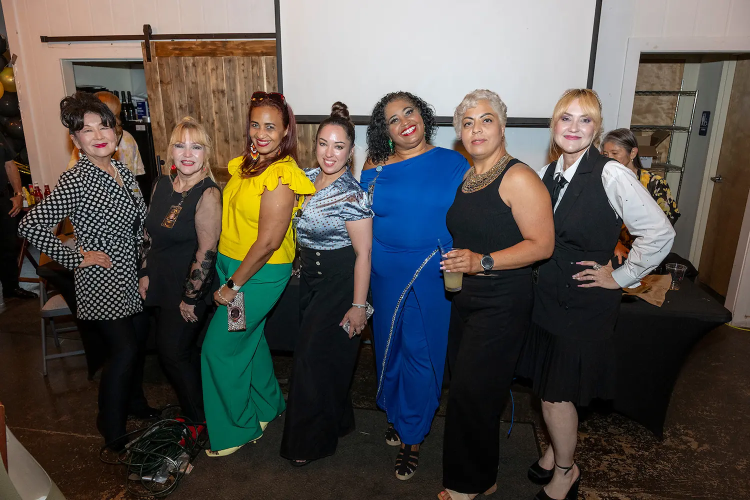 Siete mujeres permanecen juntas en un interior, vestidas con trajes elegantes y coloridos, sonriendo a la cámara. De fondo, una pantalla de proyección, paneles de madera y un ambiente festivo.