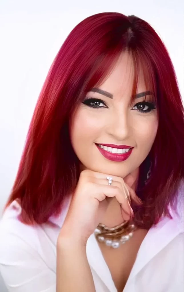 A woman with straight, shoulder-length red hair and bangs smiles confidently, resting her chin on her hand. She wears a white top, bold makeup, a necklace, and a ring on her finger. The background is softly blurred.