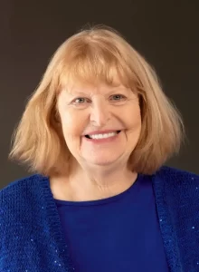 Mujer mayor sonriente con el pelo rojo claro en un top azul y un jersey de punto azul, de pie delante de un fondo oscuro liso.
