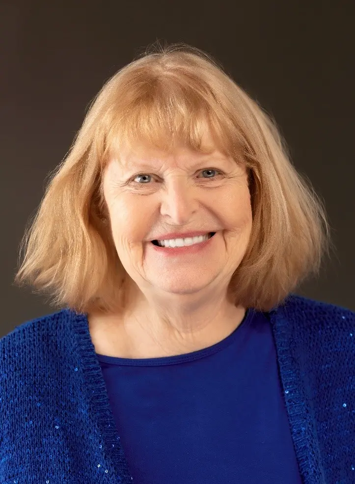 Smiling older woman with short, light reddish-blonde hair wearing a bright blue top and a blue knit cardigan, against a plain dark background.