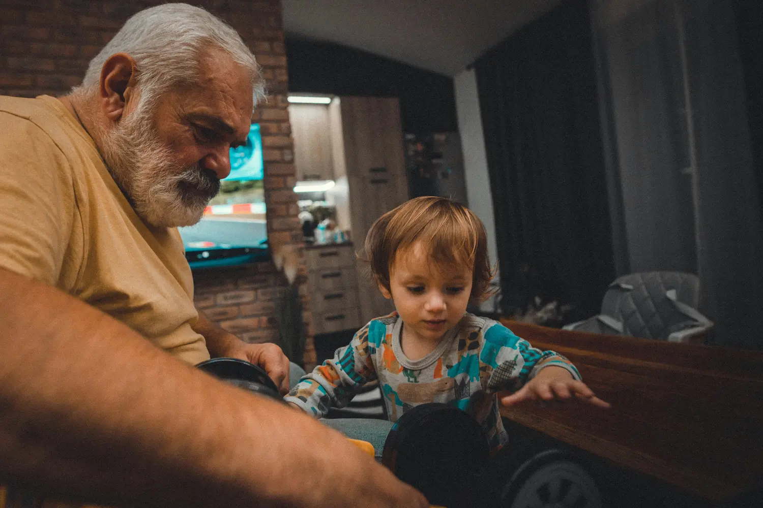 Un anciano de pelo canoso y barba se sienta a una mesa y ayuda a un niño pequeño con un coche de juguete. El niño, que lleva un pijama de colores, parece concentrado mientras alcanza el juguete. La habitación está poco iluminada y es acogedora.