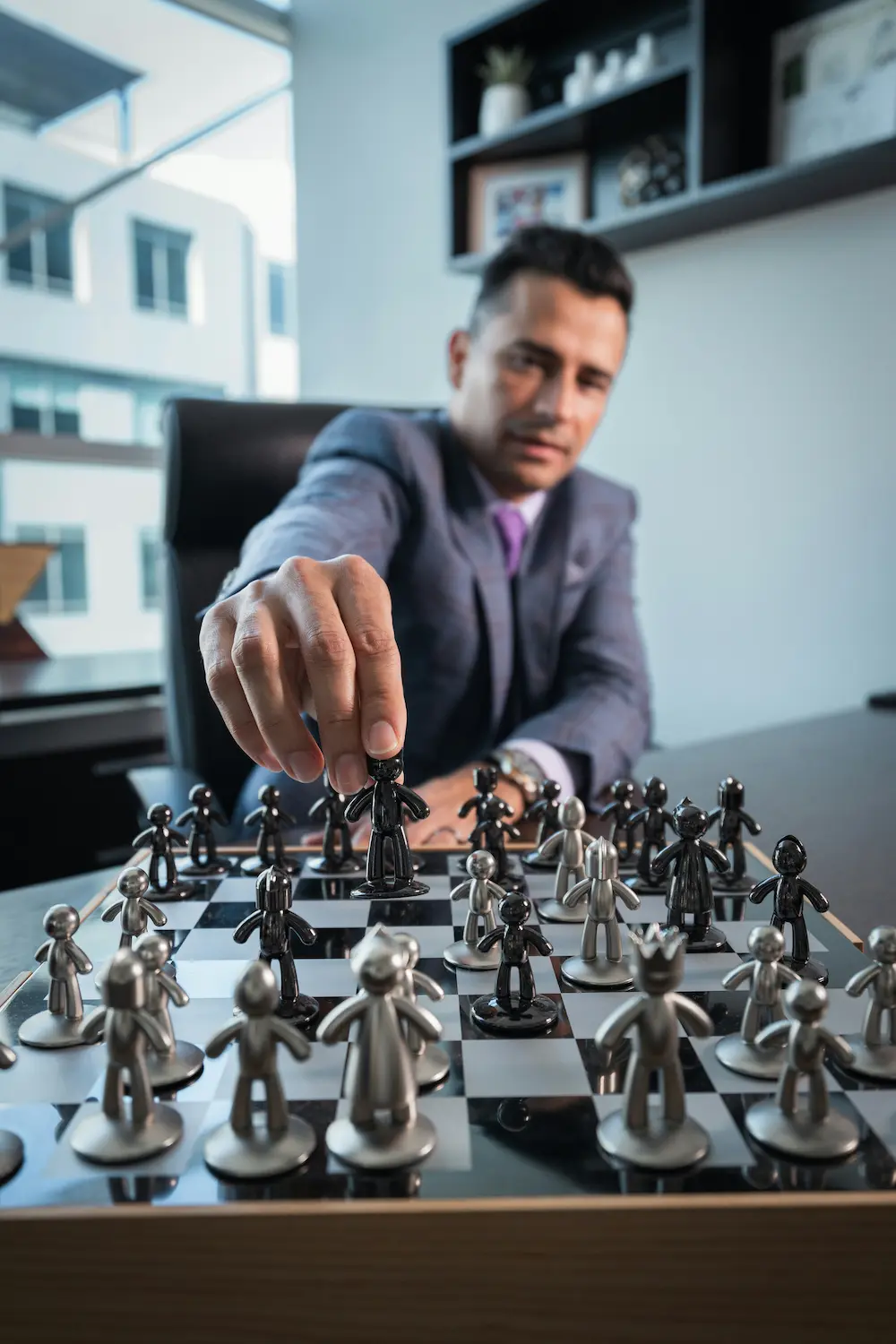 A man in a suit sits at a desk and moves a chess piece on a modern chessboard, demonstrating key leadership qualities with strategic moves, in an office setting featuring shelves and windows in the background.