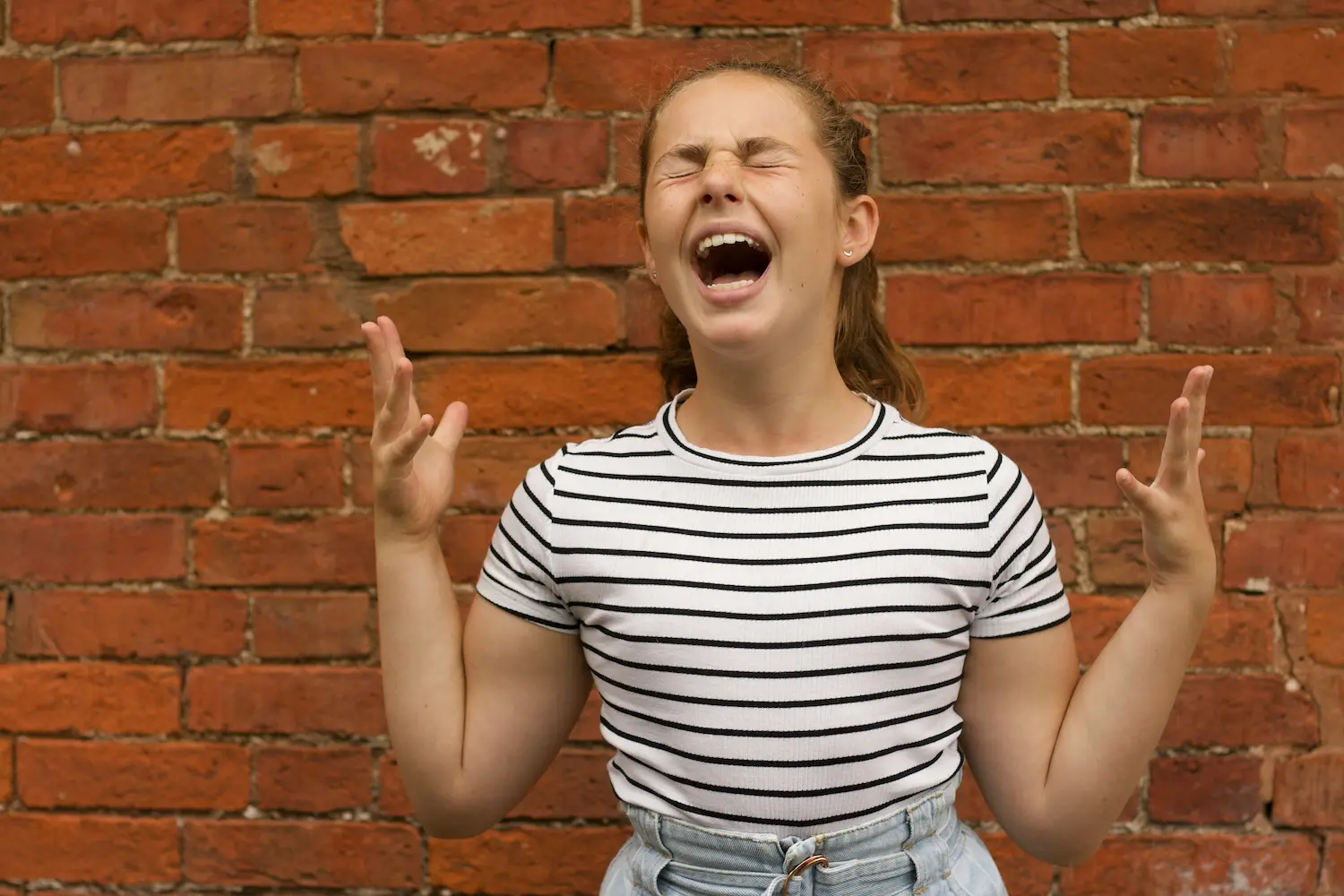 A young person in a striped shirt stands in front of a brick wall, eyes closed and mouth open wide, raising both hands in frustration or anger—as if letting out The Last Whoop.