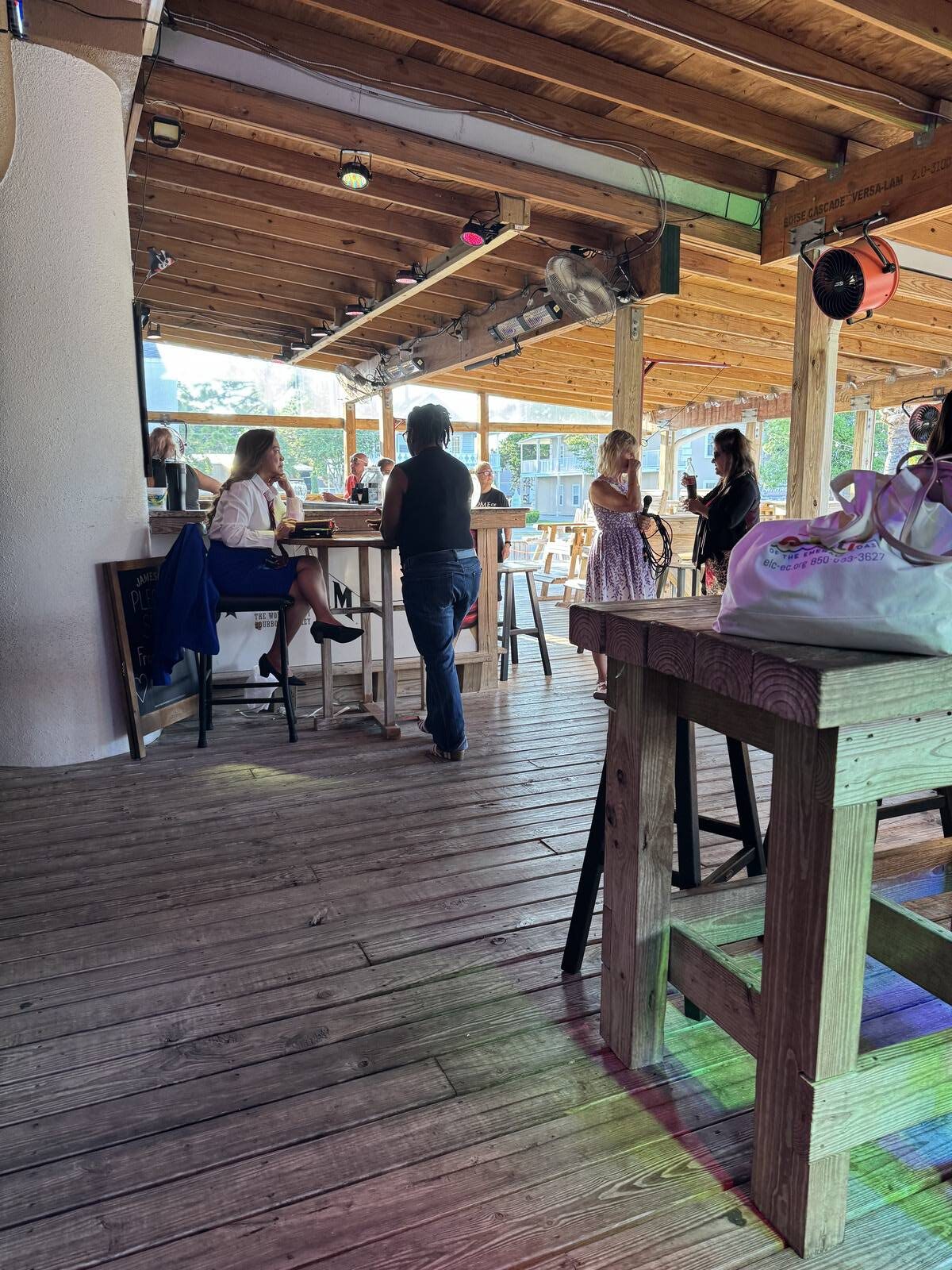 A wooden patio with several people socializing during a Hispanic Heritage Month Kick-off; some are sitting at tables while others stand and talk. Sunlight streams through open sides, and a white bag rests on a nearby wooden table in the foreground.