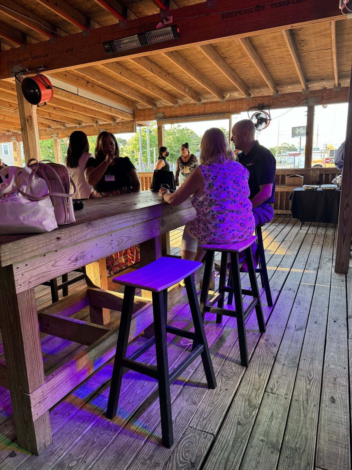 People are socializing at a wooden bar with high stools inside an open-air, wooden-roofed venue. Sunlight and purple light set the scene as guests gather for the 2025 Hispanic Heritage Month Kick-off, with a purse resting on the counter in the lively background.