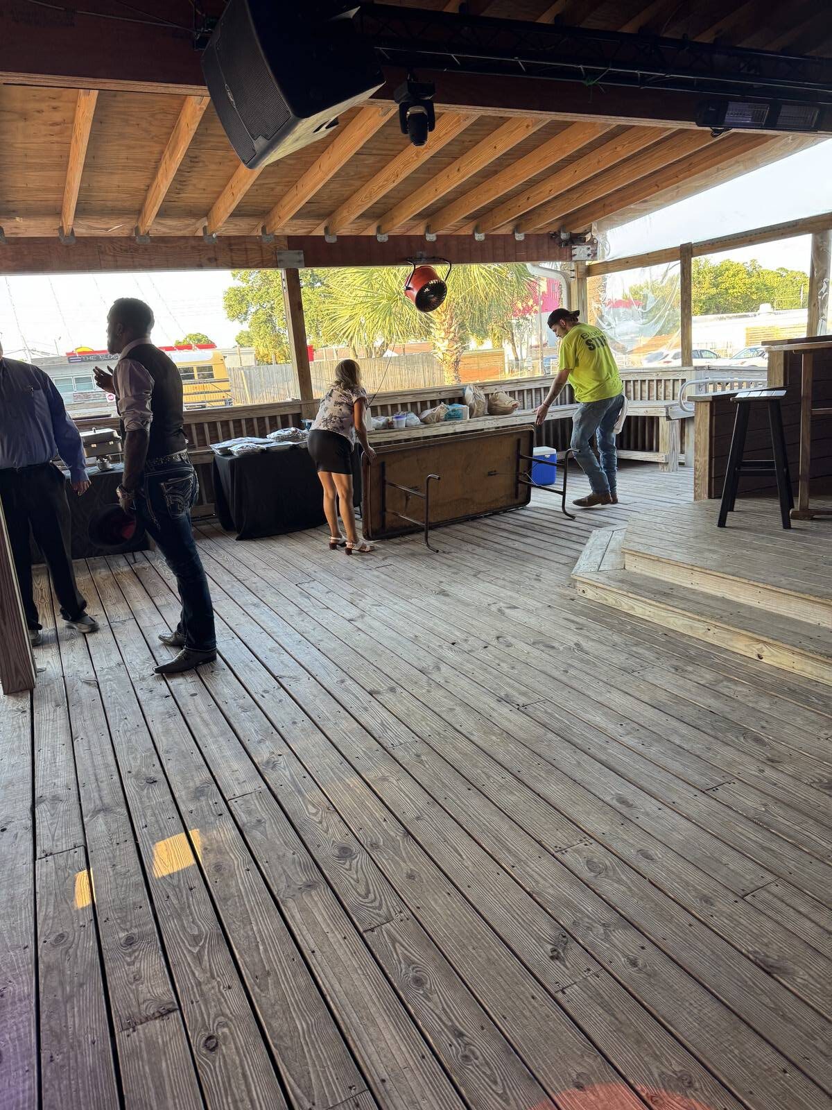 Three people move tables and chairs on a covered wooden deck with a high ceiling, preparing for the 2025 Hispanic Heritage Month kick-off. Sunlight streams through open sides, revealing trees and a blue sky outside.
