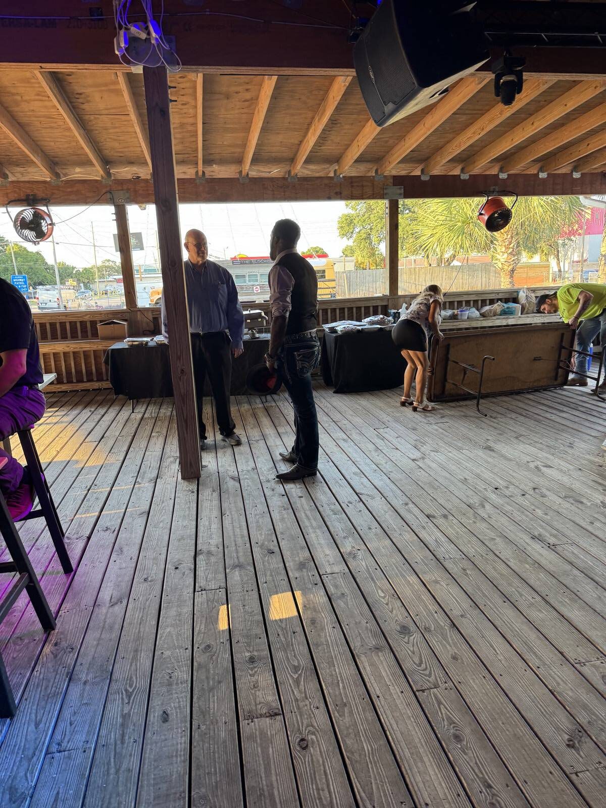 Two men stand talking on a wooden deck under a covered patio, while a woman bends over a table nearby. The scene, set outdoors with sunlight and trees, marks the Kick-off for Hispanic Heritage Month in Sept 2025.