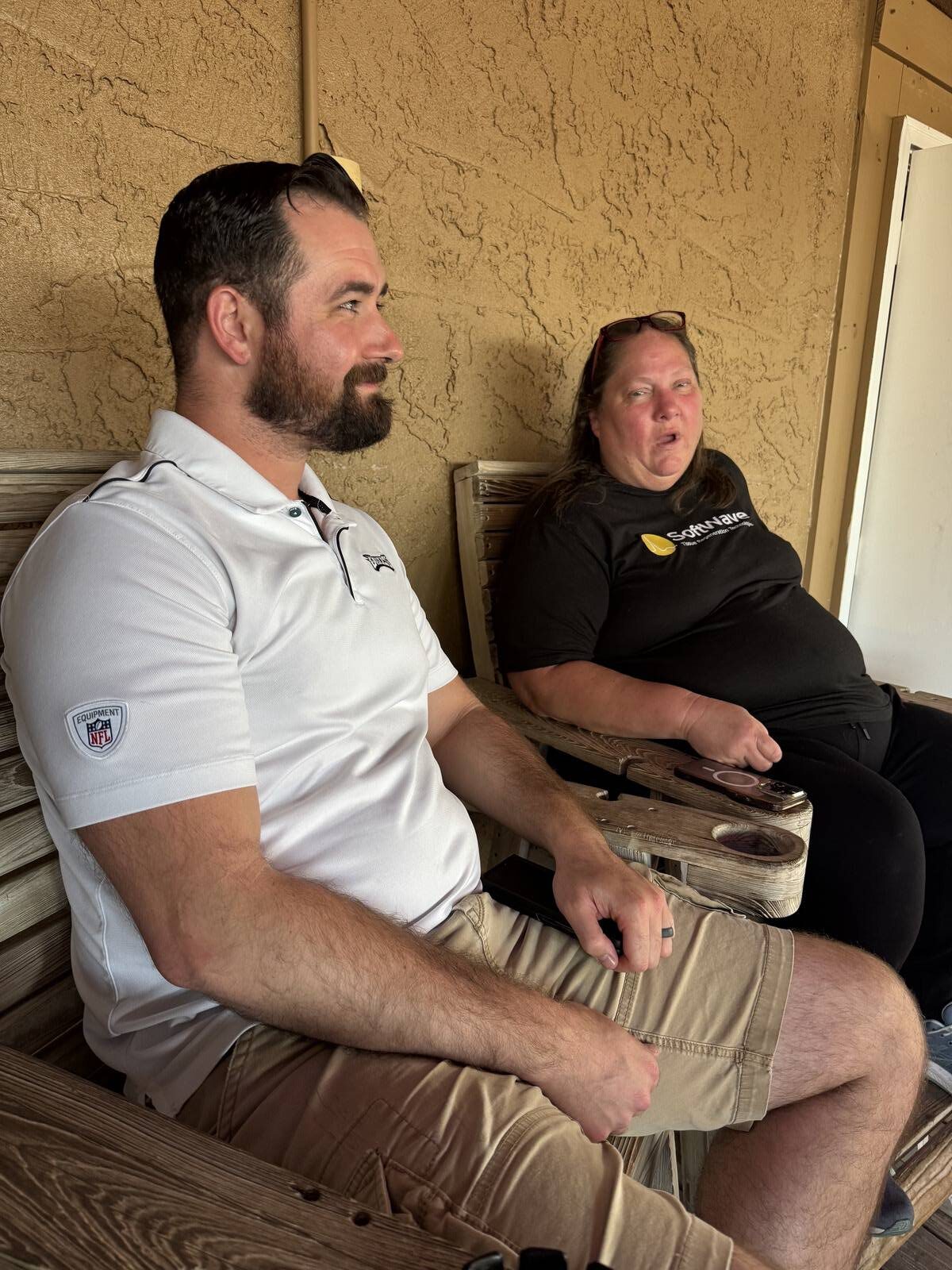 A man in a white polo and khaki shorts sits beside a woman in a black t-shirt and pants on wooden chairs against a tan textured wall, casually relaxed as they join the kick-off for Hispanic Heritage Month.